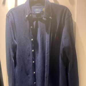 Blå långärmad skjorta från Polo Ralph Lauren - Snygg blå långärmad skjorta från Polo Ralph Lauren med klassisk krage och knappar. Den har ett broderat rött logotypmärke på bröstet. Perfekt för en stilren look.