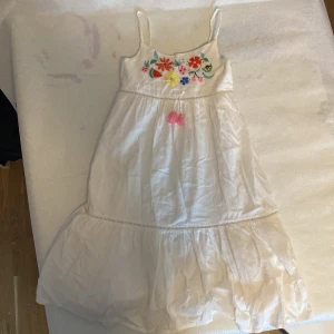 Vit klänning med broderade blommor från H&M - Charmig vit klänning från H&M med färgglada broderade blommor på bröstet och rosa tofsdetaljer. Klänningen har smala axelband och en luftig passform, perfekt för varma dagar! Klänning för barn! 