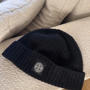 Svart ribbad mössa från Stone Island - Snygg svart ribbad mössa från Stone Island med deras ikoniska logga framtill. Perfekt för att hålla värmen under kyliga dagar. Stilren design som passar till det mesta.