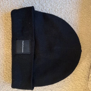 Svart stickad mössa från Peak Performance - Snygg svart stickad mössa från Peak Performance med uppvikt kant och diskret logga framtill. Perfekt för kyliga dagar och en stilren look.