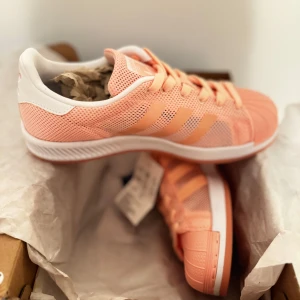 Adidas sneakers i persikofärg - Snygga Adidas sneakers i en fräsch persikofärg med vita detaljer. Skorna har en klassisk design med snörning och tre ränder på sidan. Perfekta för en sportig look.