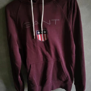 Vinröd hoodie från GANT - Säljer en snygg vinröd hoodie från GANT med ett tryck av deras logga och ett sköldemblem på bröstet. Tröjan har en klassisk känguruficka och justerbar huva med vita snören. Perfekt för en avslappnad stil.