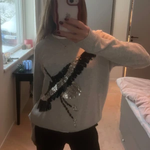 Grå tröja med paljettdetaljer - Säljer en snygg grå tröja med glittriga paljettdetaljer i svart och silver framtill. Tröjan har långa ärmar och en bekväm passform. Perfekt för att liva upp garderoben med lite glitter!