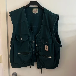 Grön väst - Snygg grön väst med flera praktiska fickor och dragkedjor. Västen har en cool design med knappar och justerbara snören i nederkant. Verkligen supercool
