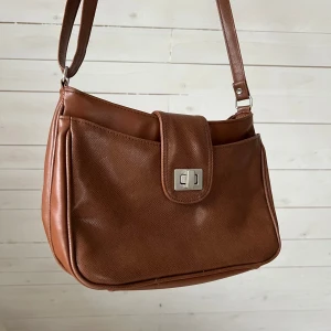 Brun axelväska i skinn - Snygg brun axelväska med skinnlock och spänne. Väskan har en justerbar axelrem och en praktisk storlek för vardag. 