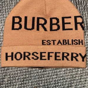 Burberry mössa - Snygg beige mössa från Burberry med svart text. Mössan har en uppvikt kant och texten 'BURBERRY ESTABLISH HORSEFERRY ROAD SW1' i svart. Perfekt för att hålla sig varm med stil. Obs skriv innan köp