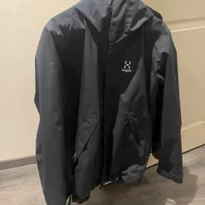 Haglöfs - Snygg mörkblå regnjacka från Haglöfs med huva och praktiska fickor. Perfekt för regniga dagar och håller dig torr och bekväm. Jackan har en stilren design med diskret logga på bröstet.
