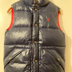 Vändbar dunväst från Polo Ralph Lauren - Snygg vändbar dunväst för barn från Polo Ralph Lauren i marinblått och rött. Storlek 10-12 år. Västen har knappar och den klassiska  loggan på bröstet. Perfekt för att hålla värmen med stil. Använd ett par gånger under en säsong. 