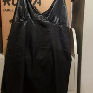 Svart klänning med spetsdetaljer - Elegant svart klänning med spetsdetaljer vid axelbanden. Klänningen har en v-ringad design och är tillverkad i ett glansigt material som ger en lyxig känsla.