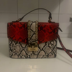 Röd och beige axelväska med ormmönster - Snygg axelväska med ormmönster i rött och beige. Väskan har en guldfärgad kedjedetalj och ett praktiskt lås framtill. Perfekt för att ge din outfit en trendig touch.