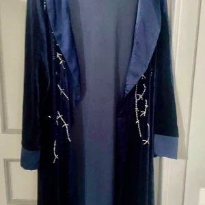 Svart sammet Abaya med pärlor - Elegant svart Abaya i sammet med vackra pärldetaljer längs framsidan. Den har långa ärmar och en klassisk design som ger en lyxig känsla. Perfekt för speciella tillfällen.
