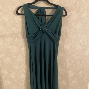 Mörkgrön klänning från H&M - Elegant mörkgrön klänning från H&M med en stilren design. Klänningen har en knytning runt halsen och en dekorativ ringdetalj framtill. Perfekt för speciella tillfällen.😊