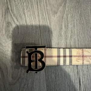 Burberry bälte - Snyggt Burberry’s bälte med ett stort monogramspänne. Ena sidan har ett klassiskt rutigt mönster i beige, svart och röd, medan den andra sidan är svart med diskreta rutor. Perfekt för att ge en stilren touch till din outfit.