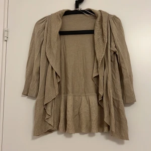 Beige kofta från Valleygirl - Säljer en tunn beige kofta från Valleygirl med volangdetaljer. Koftan har trekvartsärmar och en lös passform, perfekt för lager-på-lager under hösten. Den är tillverkad i ett mjukt stickat material.