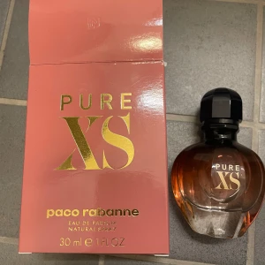 Pure XS Eau de Parfum från Paco Rabanne - Säljer en elegant och sensuell Pure XS Eau de Parfum från Paco Rabanne. Flaskan är i en stilren design med en svart kork och en gradient av rosa till bärnstensfärg. Perfekt för den som vill ha en doft som utstrålar lyx och självsäkerhet.