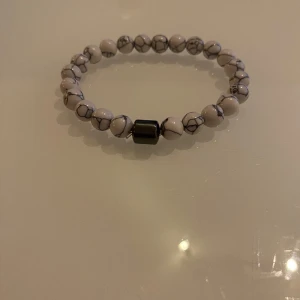 Vitt pärlarmband - Snyggt armband med vita pärlor med grå ådring och en större mörkgrå pärla i mitten. Perfekt för att ge en stilren touch till din outfit.