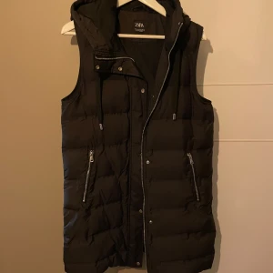 Svart dunväst från Zara - Snygg svart dunväst från Zara med dragkedja och knappar. Perfekt för kyliga dagar med sin quiltade design och praktiska fickor med dragkedja. Huvan ger extra skydd mot väder och vind. Använd fåtal gånger 
