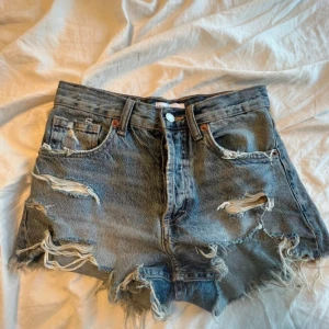 Blå jeansshorts med slitningar - Säljer ett par coola blå jeansshorts med slitningar och fransiga kanter. De har en klassisk femficksdesign och knappgylf. Perfekta för en avslappnad sommarlook!
