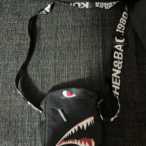 Svart axelväska med hajmotiv - Cool svart axelväska med ett hajmotiv på framsidan och text på axelremmen. Väskan har en dragkedja för säker förvaring och är perfekt. Är inte använd. 