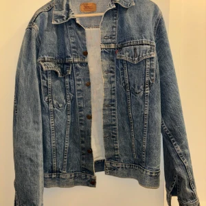 Jeansjacka från Levi's med broderad rygg - Snygg jeansjacka från Levi's med en unik broderad design på ryggen som säger 'Bill the Cat Lives'. Jackan har knappar framtill och två bröstfickor med lock. Perfekt för en laidback stil. Jackan är vintage och passar M eller stor Small
