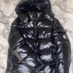 Moncler Maya - Snygg svart pufferjacka från Moncler med glansig finish och huva. Jackan har en dragkedja framtill och en ficka med knappdetalj på ärmen. Perfekt för kyliga dagar.