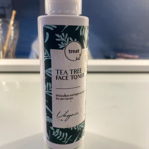 Tea Tree Face Toner - Säljer en Tea Tree Face Toner från märket Treat. Denna toner återfuktar och hjälper till att reparera hudbarriären. Den är vegansk och perfekt för daglig användning. Flaskan är vit med en grön etikett som har ett bladmotiv.