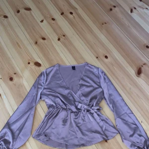 Lila omlottopp från Shein - Säljer en superfin lila omlottopp från Shein. Den är gjord i ett glansigt material som känns som satin och har långa ärmar. Toppen har en knytning i midjan som ger en snygg passform. Perfekt för fest eller en utekväll! 💜