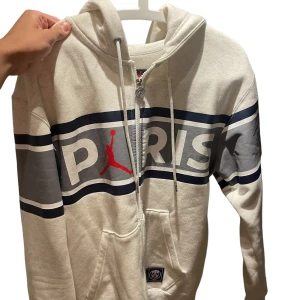 Vit hoodie från Jordan x PSG - Säljer en snygg vit hoodie från Jordan x PSG. Den har en dragkedja framtill och en stor ficka på magen. Hoodien har ett coolt tryck med texten 'PARIS' och Jordan-loggan i rött. Perfekt för höst och vinter!