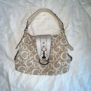 Beige och vit axelväska med mönster - Säljer en snygg beige och vit Guess axelväska med ett coolt mönster. Väskan har en justerbar axelrem och en stor spänne framtill. Perfekt för vardagsbruk eller en dag på stan. Väskan är i bra skick och redo för en ny ägare!