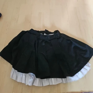 Svart kjol med vit underkjol - Säljer en svart kjol med en vit underkjol som sticker fram längst ner. Kjolen är kort och har en volangdetalj som ger den en söt och lekfull look. Perfekt för fest eller en dag ute på stan!