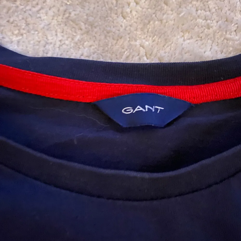 Säljer en snygg mörk blåa t-shirt från GANT. Den har en vit och röd randig detalj över bröstet med GANT-loggan i mitten. T-shirten är i bomull och har en normal passform. Perfekt för vardagsbruk eller en avslappnad dag ute. Skicket är bra men en liten tråd hänger från den. Passa på att fynda! 😎. T-paidat.