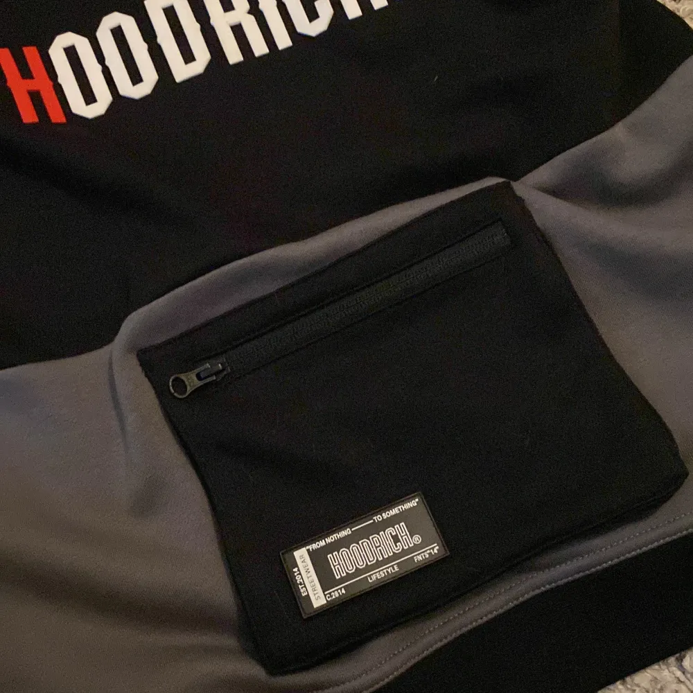 Säljer en snygg hoodie från Hoodrich i färgerna svart, grå och röd. Den har en stor ficka med dragkedja på framsidan och texten 'Hoodrich' tryckt både fram och bak. Perfekt för höst och vinter!. Tröjor & Koftor.