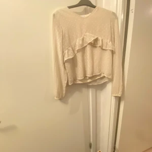 Beige blus med volangdetaljer - Säljer en söt beige blus med volangdetaljer framtill. Blusen har långa ärmar och en v-ringad hals. Perfekt för både vardag och festliga tillfällen. Passar bra till jeans eller kjol för en chic look!