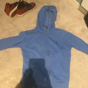 Blå hoodie från Polo Ralph Lauren - Säljer en snygg blå hoodie från Polo Ralph Lauren. Den har en dragkedja framtill och en klassisk logga på bröstet. Perfekt för en avslappnad stil och passar bra till både höst och vår. Skön och stilren, ett måste i garderoben! 💙 den är i riktigt bra skick, köpt från Nk för ca 1200 storlek är 14-16 men skulle säga att den passar 145-160