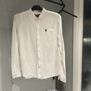 Vit skjorta från Lyle & Scott - Säljer en stilren vit skjorta från Lyle & Scott i storlek 15/16 år. Skjortan har långa ärmar och en klassisk krage med knappar. Den är i bomull och har en broderad logga på bröstfickan. Perfekt för både vardag och fest!