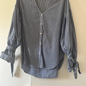 Randig blus med puffärm - En söt v-ringad blus med puffärm och knyt runt handlederna. Den är blå/vit randig och är lite längre bak än fram. I nyskick och endast använd fåtal gånger