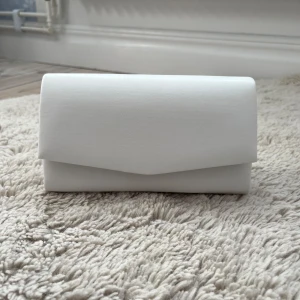 Vit kuvertväska - Snygg och stilren vit kuvertväska i syntetmaterial som endast är använd 1 gång till balen. Den har lite små defekter (skriv för bilder) men annars i jätte bra skick💕 (tillkommer även ett kedjeband så kan bäras på olika sätt).                                          Längden - 18 cm, bredden - ca 10.5 cm