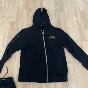 Säljer en snygg svart hoodie från Boss med dragkedja och huva. Den har coola detaljer på ärmarna och Boss-loggan på bröstet. Perfekt för en avslappnad stil eller träning. Passar både höst och vår.