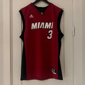 Röd basketlinne från Adidas - Säljer en röd basketlinne från Adidas med Miami-tryck och nummer 3 på framsidan. På baksidan står det 'Wade' med samma nummer. Linnet är i mycket bra skick och perfekt för basketfans. Det är ärmlöst och har svarta sidopaneler för en snygg kontrast.