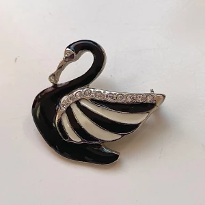 Elegant svanbrosch i svart och vitt - Säljer en superfin brosch i form av en svan. Den är svart och vit med glittrande stenar på vingarna. Perfekt för att piffa upp en outfit till fest eller bal. Broschen är cirka 4 cm lång och har en säkerhetsnål på baksidan för enkel fastsättning.