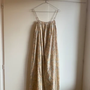 Beige långklänning med blommigt mönster - Säljer en superfin beige långklänning med ett diskret blommigt mönster i blått och brunt. Klänningen har smala axelband, en luftig passform och öppen rygg med knytning. Den är i köpt second hand men är som ny!🩷