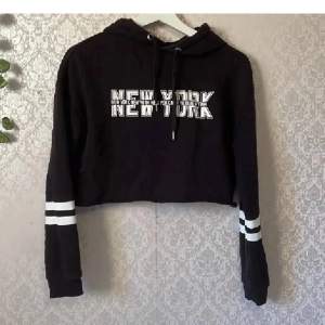 Säljer en svart croppad hoodie med texten 'New York' i vitt på bröstet. Den har långa ärmar med vita ränder vid ärmsluten och en justerbar huva med snören. 