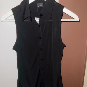 Collar Tanktop Gina tricot S, med prislapp på - Snygg svart ärmlös topp med krage och knappar framtill. Perfekt för både vardag och festliga tillfällen. Tillverkad i ett mjukt och bekvämt material som ger en stilren look. Passar bra till både kjol och byxor. Tröjan är helt oanvänd eftersom storleken är liten för mig🥰