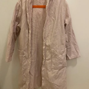Ljusrosa badrock från H&M - Säljer en badrock från H&M i storlek 122/128 för barn. Tillverkad i bomull med huva och knytskärp i midjan. Perfekt för att hålla sig varm efter badet eller på morgonen. Två praktiska fickor framtill.