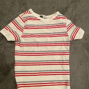 Randig t-shirt från H&M - Säljer en fräsch randig t-shirt från H&M i vitt med röda och blå ränder. Perfekt för en avslappnad stil. T-shirten är kortärmad och gjord i mjuk bomull. Passar bra till både jeans och shorts!