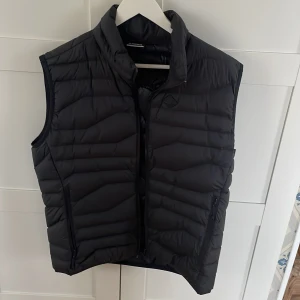 Svart dunväst från J.Lindeberg - Säljer en snygg svart dunväst från J.Lindeberg i mycket bra skick. Västen har en dragkedja framtill och en stilren design med ett diskret märke på bröstet. Perfekt för höst och vår när du vill hålla värmenen utan att kompromissa med stilen. Storlek XL men passar L. Nypris 2000kr.