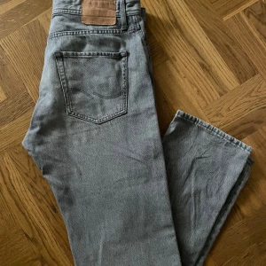 Grå jeans från Jack & Jones - Säljer ett par grå jeans från Jack & Jones i modellen Relaxed Chris. De har en avslappnad passform och är perfekta för en casual look. Jeansen har en klassisk femficksdesign och knappar i silver. Storlek 30/30