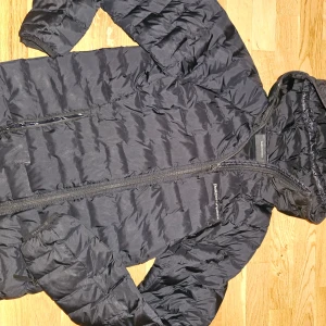 Svart dunjacka från Peak Performance - Säljer en svart dunjacka från Peak Performance i mycket bra skick. Jackan har en praktisk dragkedja och är perfekt för kalla dagar. Den är långärmad och har en skön huva för extra värme. Perfekt för både vår, höst och vinter! Märkesjacka, kvalitejacka, superfintskick. 