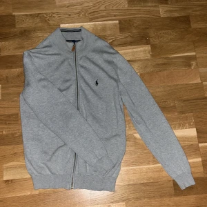 Grå kofta från Polo Ralph Lauren - Säljer en stilren grå kofta från Polo Ralph Lauren i storlek M kan även passa S. Den har en dragkedja framtill och är tillverkad i mjukt material. Perfekt .för både höst och vår. Den klassiska loggan är broderad på bröstet. Kontakta via övriga frågor.