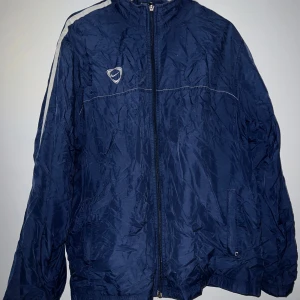 Blå tunnare jacka från Nike - Säljer en blå tunnare jacka från Nike. Tyvärr är den lite skrynklig just nu då den legat i en påse ett tag. Jackan har en dragkedja framtill och en vit rand längs ärmarna. Med en normal passform och långärmade ärmar. Den har sina skavanker och därav säljs den billigt, bifogat bilder på skavankerna.